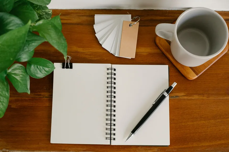 An open notebook with a pen, beside a mug and a small potted plant on a desk.